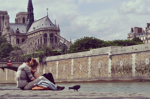 Venku na ulici na betonu sedí mladý pár, kolem nich jsou holubi a opodál katedrála